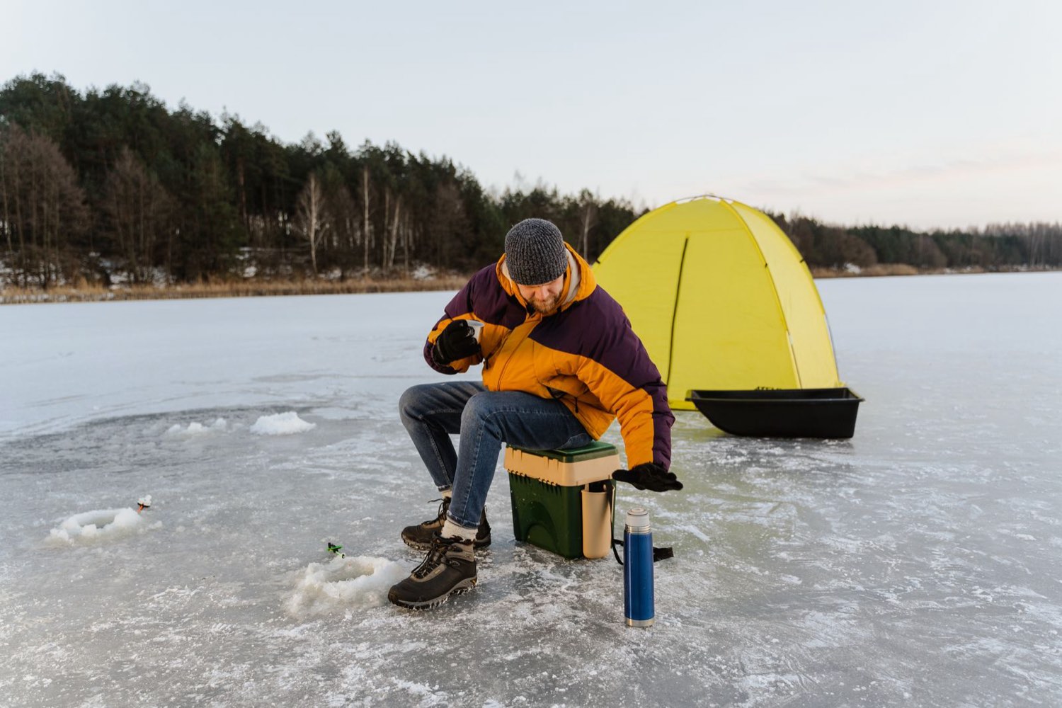 Angler enjoying a hot drink beside a bright tent on a frozen lake